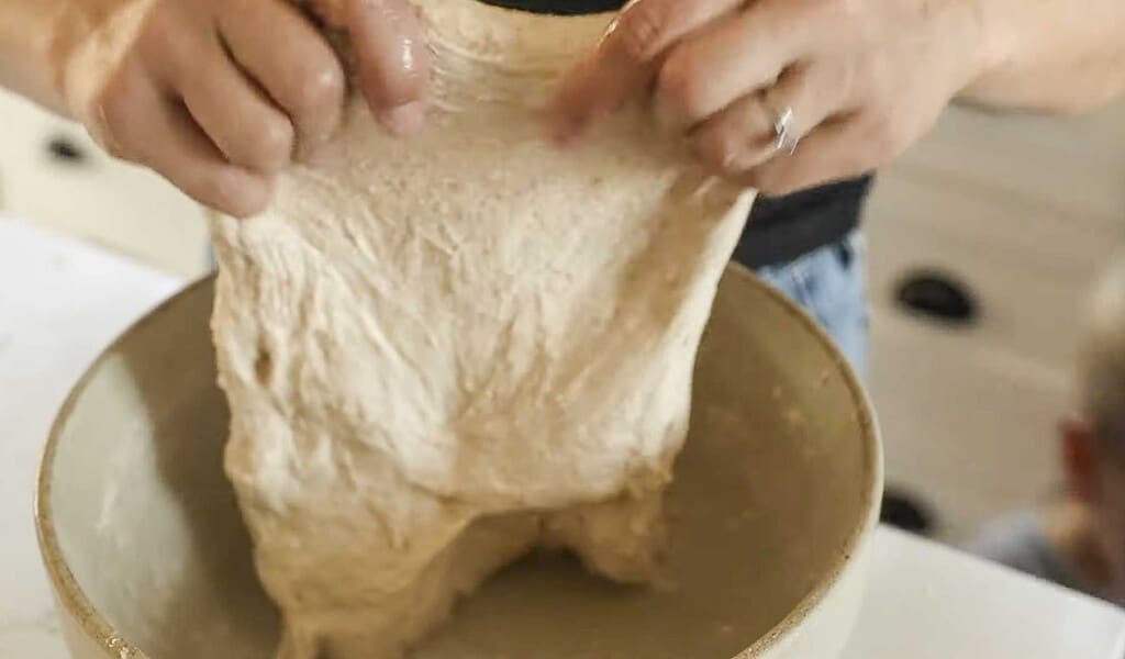 hands holding bread dough and pulling it up out of a stoneware bowl