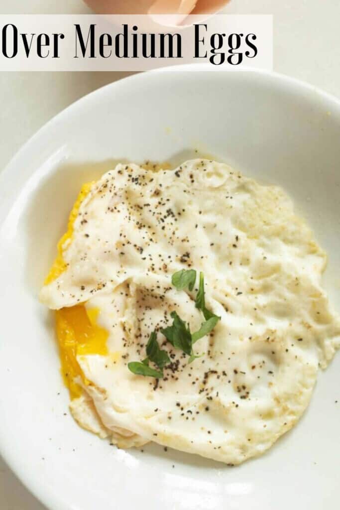 a white plate with an over medium egg topped with fresh basil on a white countertop with a cracked egg to the left