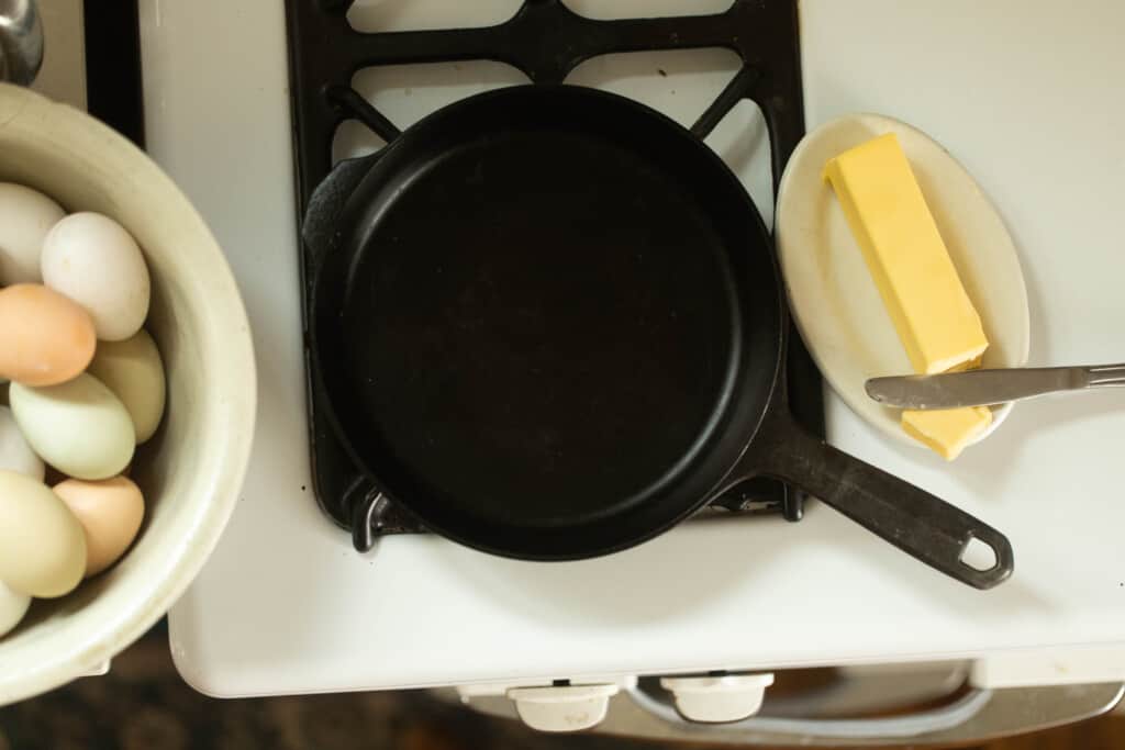 cast iron skillet on a stove with a plate of butter with a knife to the right and a large bowl of eggs to the left