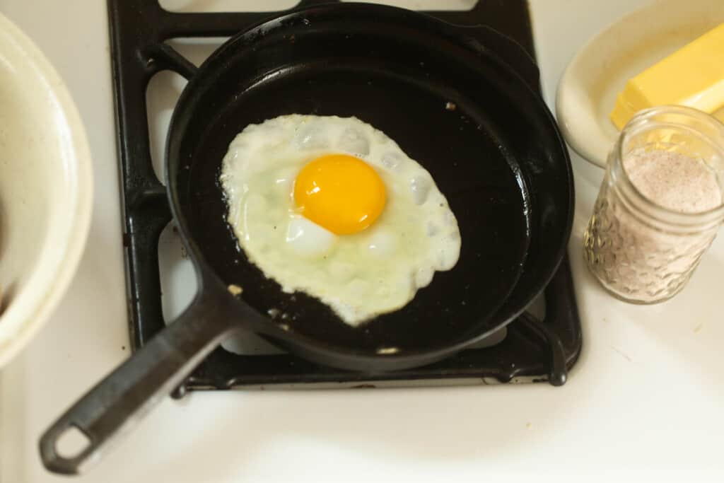egg cooking in a cast iron skillet on a white stove with butter on a plate to the right