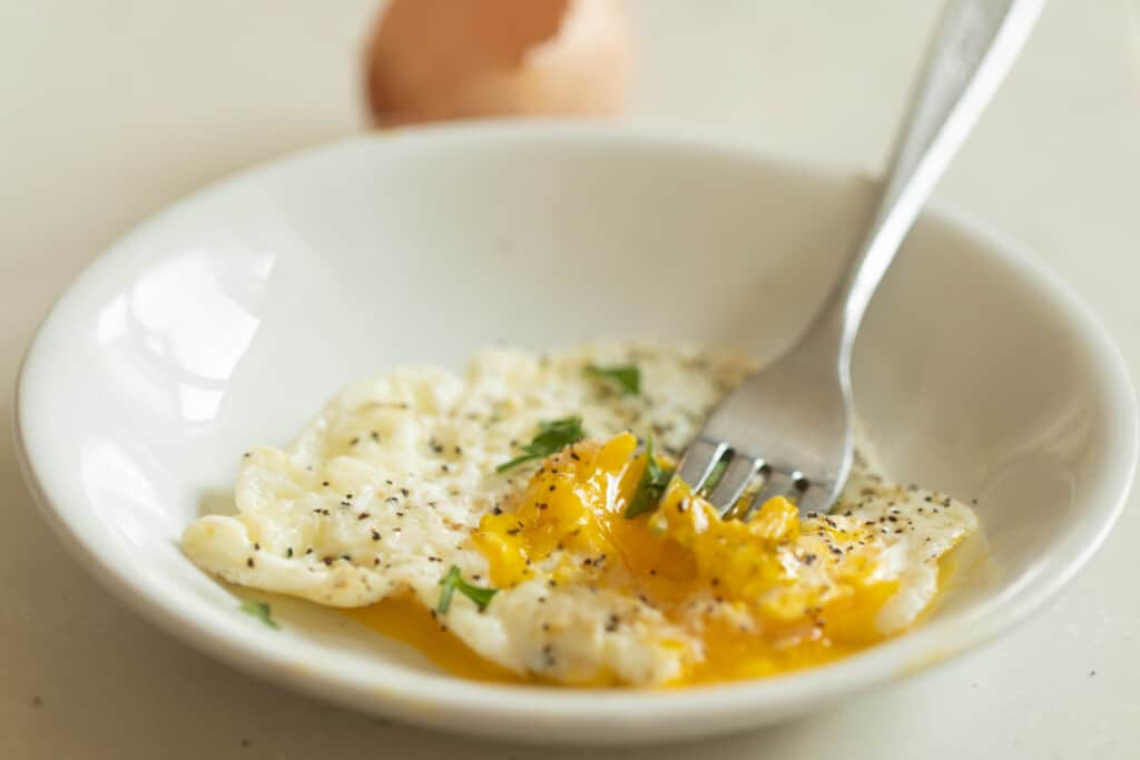 a fork in the runny yolk of an over-easy egg in a white bowl with a egg in the background