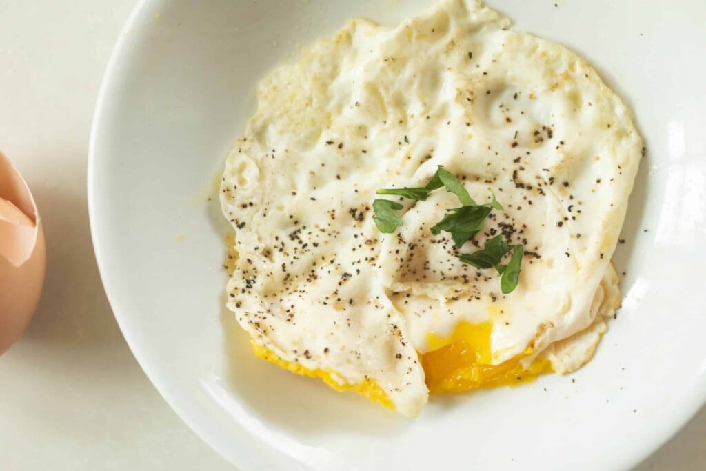 overhead photo of an over medium egg topped with fresh herbs on a white bowl on a white countertop with a cracked egg in the back