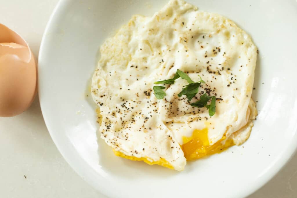 a white plate with an over medium egg topped with fresh basil on a white countertop with a cracked egg to the left