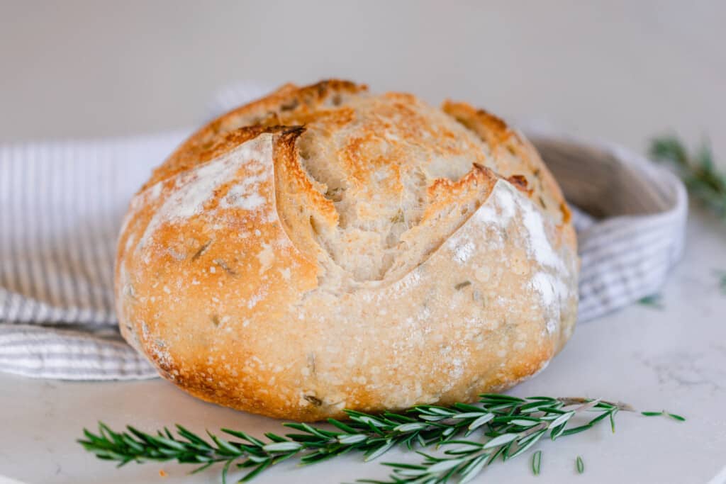 loaf of rosemary sourdough bread on a white countertop with a sprig of rosemary in the front