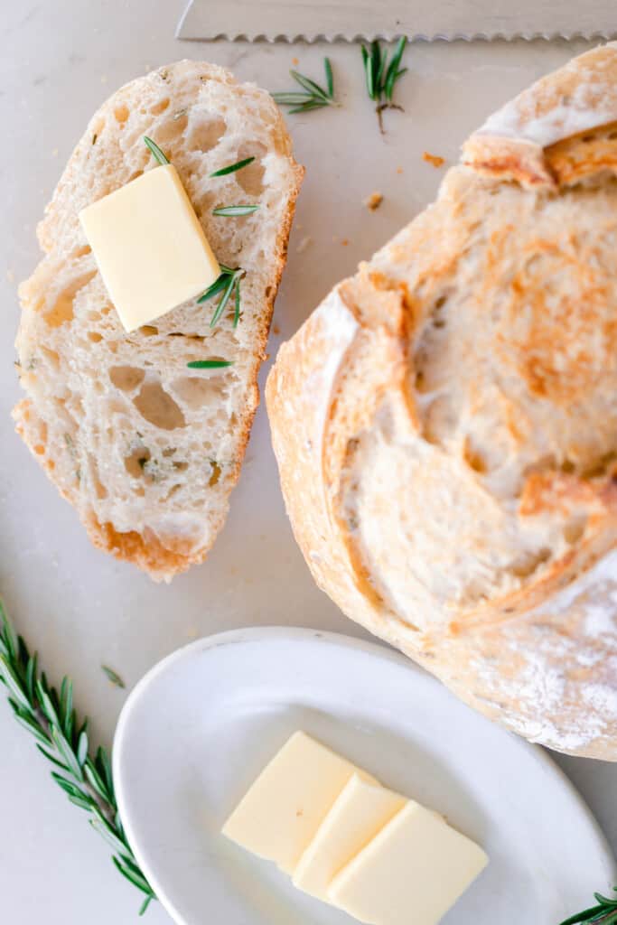 rosemary sourdough boule with a slice of bread to the left topped with a pat of butter. A small dish of butter is in front of the loaf