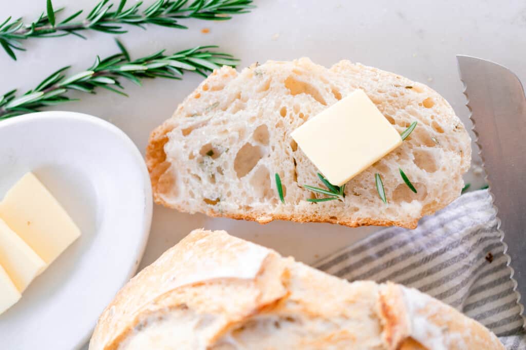 slice of sourdough rosemary bread with a pat of butter on a white countertop with a white dish with butter and sprigs of rosemary to the left