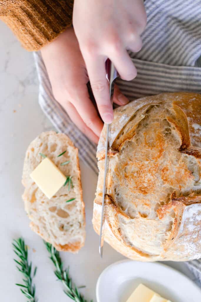 hand holding a knife and slicing a slice of rosemary sourdough bread off of a loaf of bread. Another slice is to the left topped with a pat of butter