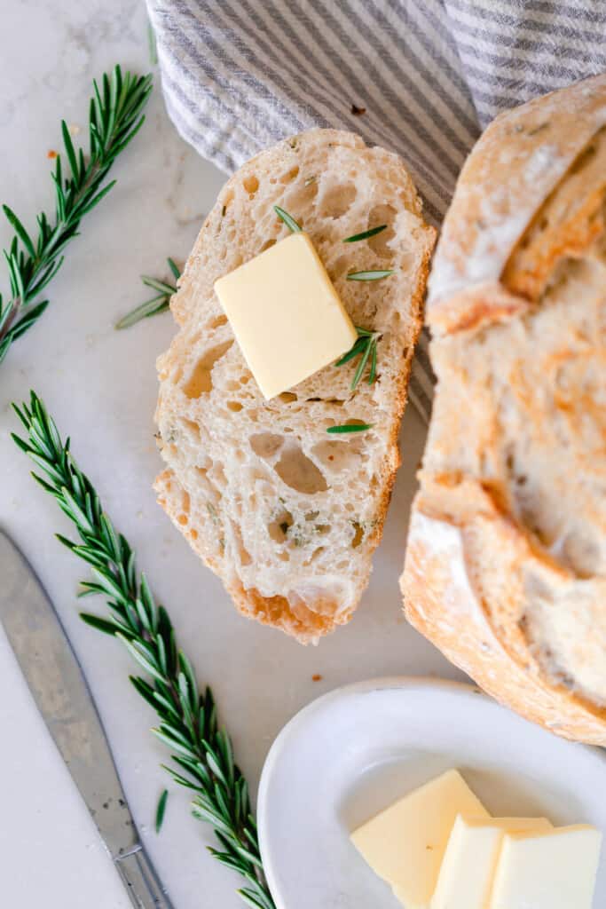 overhead photo of a loaf of rosemary sourdough bread with a slice off of it and resting to the left side of the loaf. Sprigs of rosemary are placed around the bread
