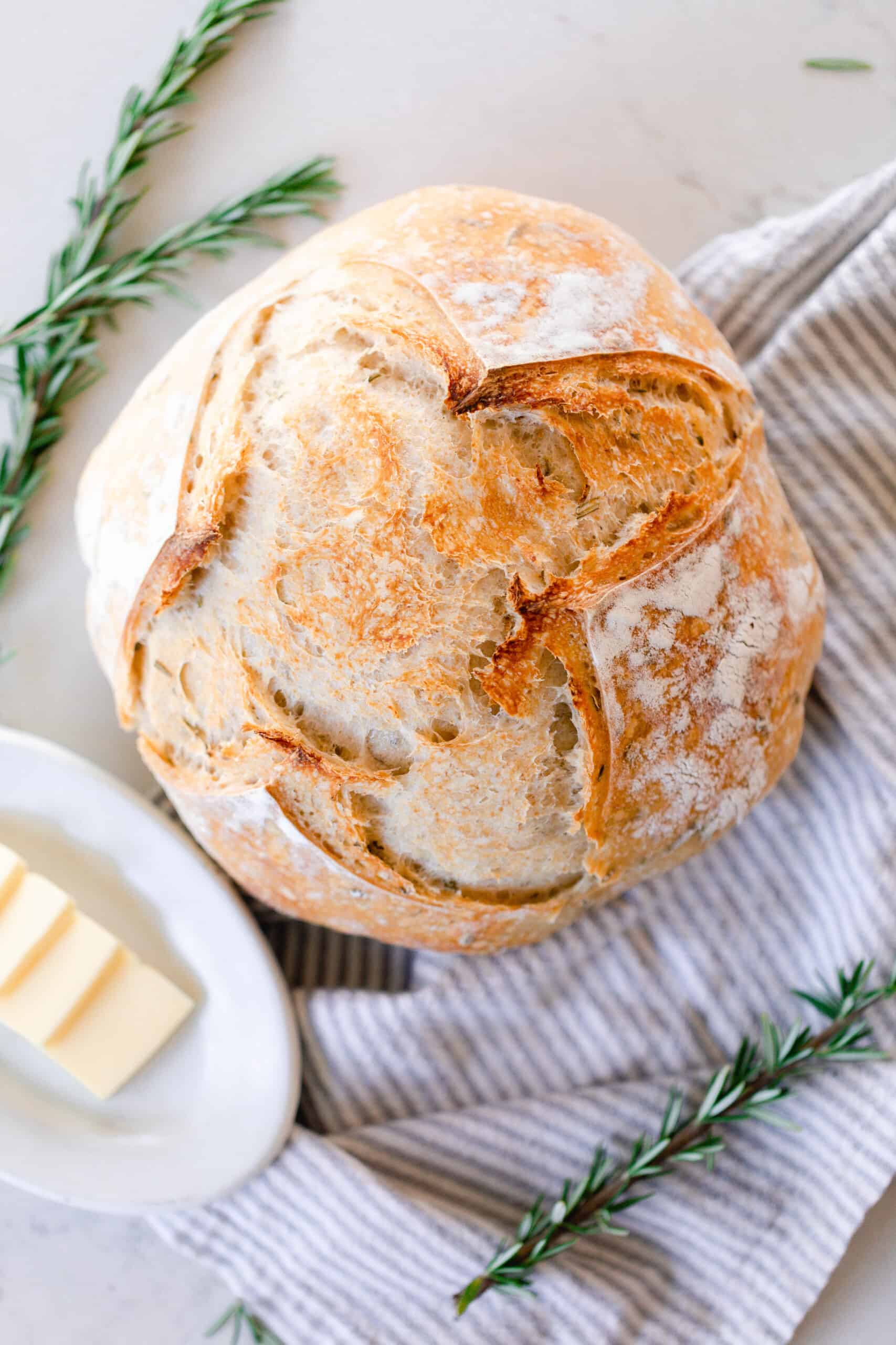 overhead photo of a loaf of crust rosemary sourdough bread on a gray and white towel. With fresh springs of rosemary around the loaf