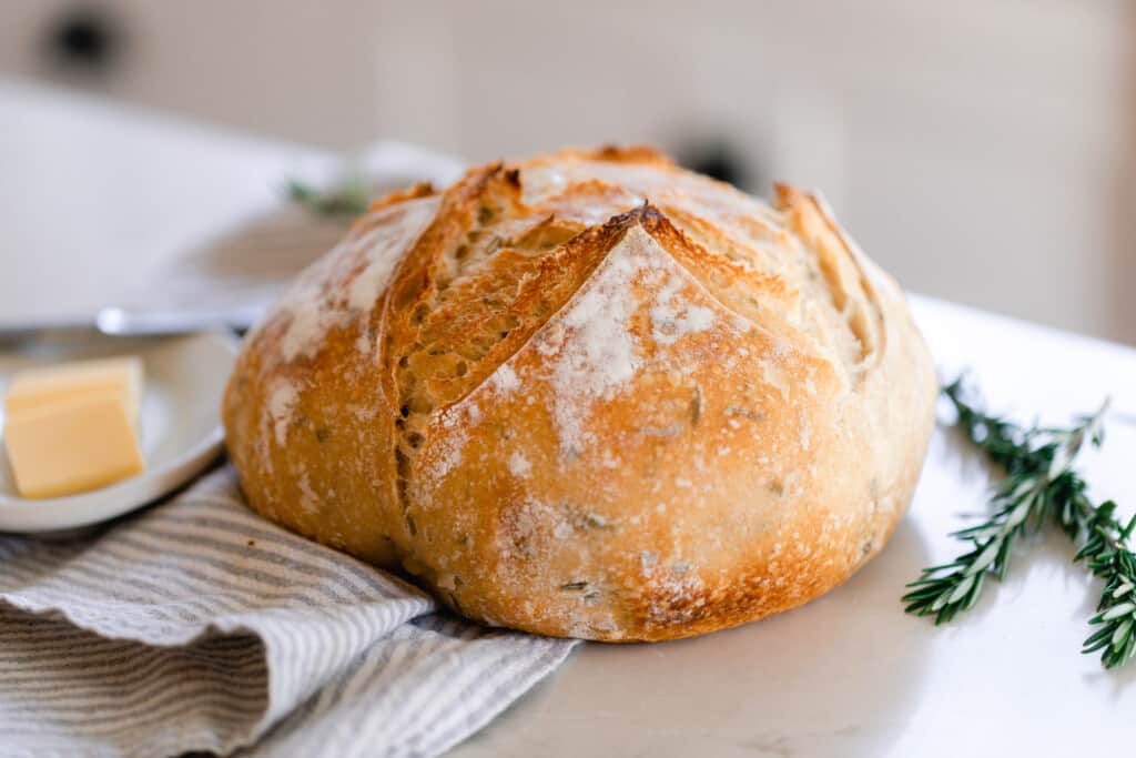 round boule of rosemary sourdough bread on a gray and white stripped towel white countertop. Fresh sprigs of rosemary are to the right of the bread.