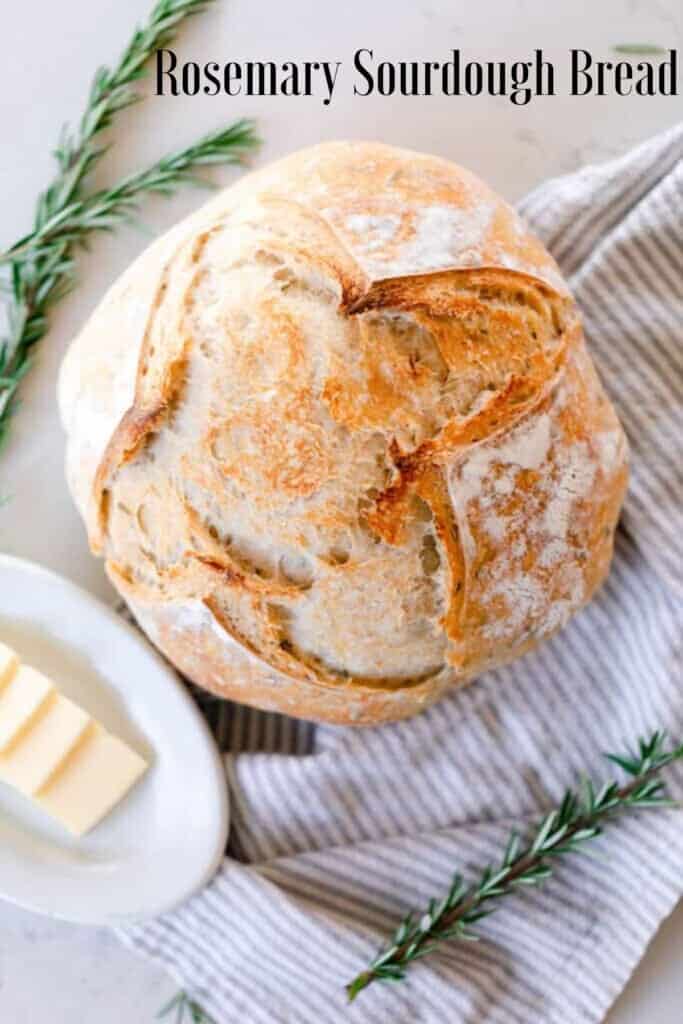 overhead photo of a loaf of crust rosemary sourdough bread on a gray and white towel. With fresh springs of rosemary around the loaf