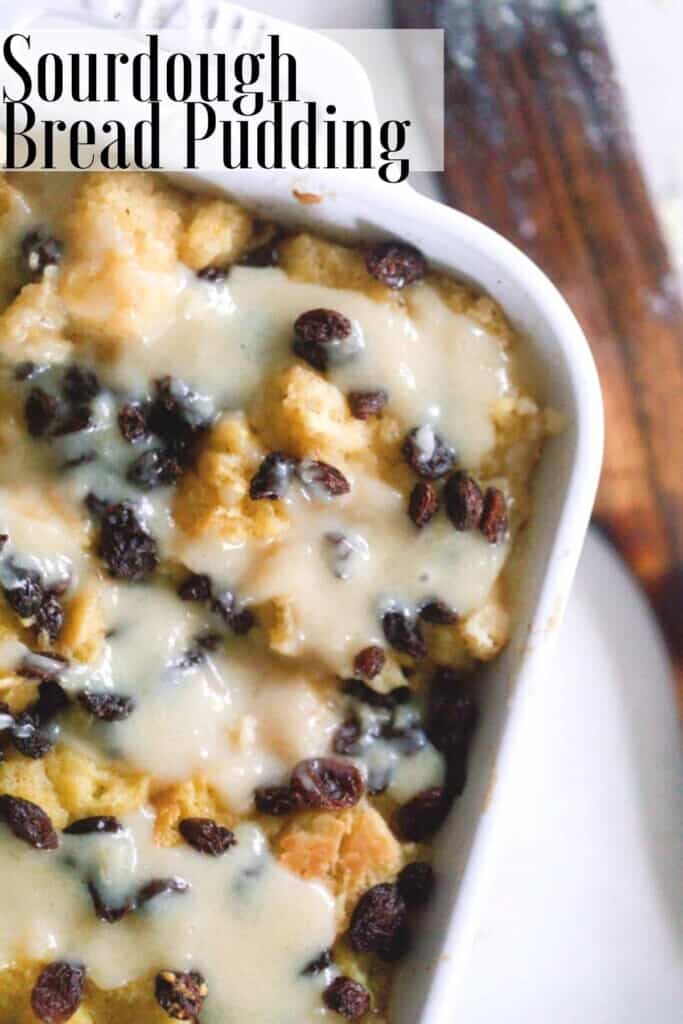 overhead photo of sourdough bread pudding topped with raisins and a creamy sauce in a white baking dish on a white countertop with a wooden spatula to the right