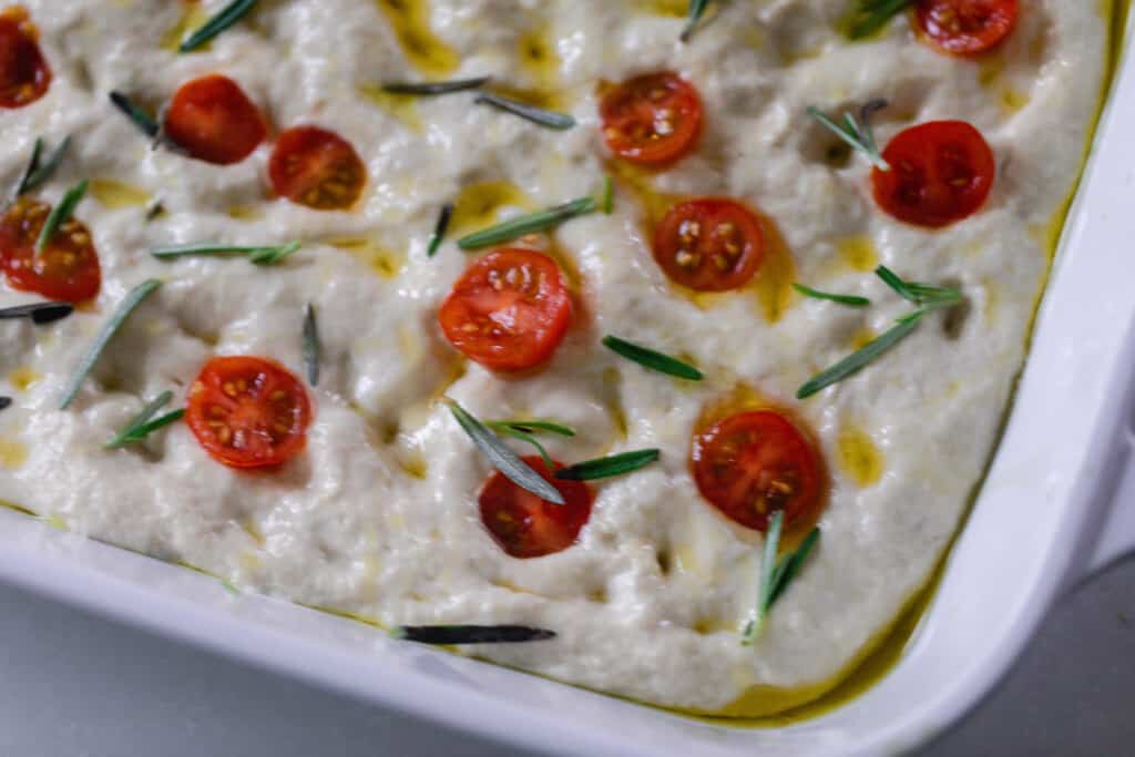 close up of sourdough focaccia dough topped with sliced cherry tomatoes and fresh rosemary in a white baking dish