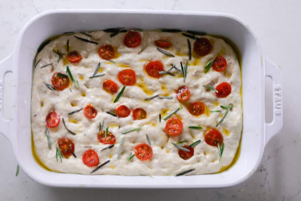 overhead photo of sourdough focaccia dough topped with tomatoes and fresh rosemary in a white baking dish