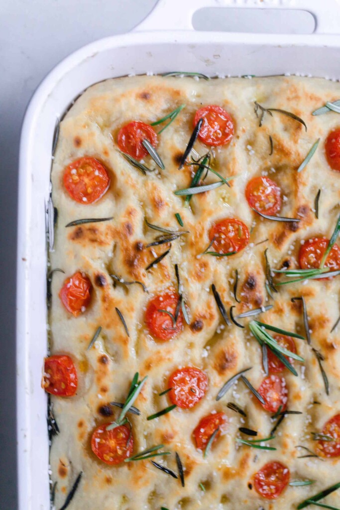 overhead photo of 1/4 of a white baking dish with sourdough focaccia topped with tomatoes and fresh rosemary