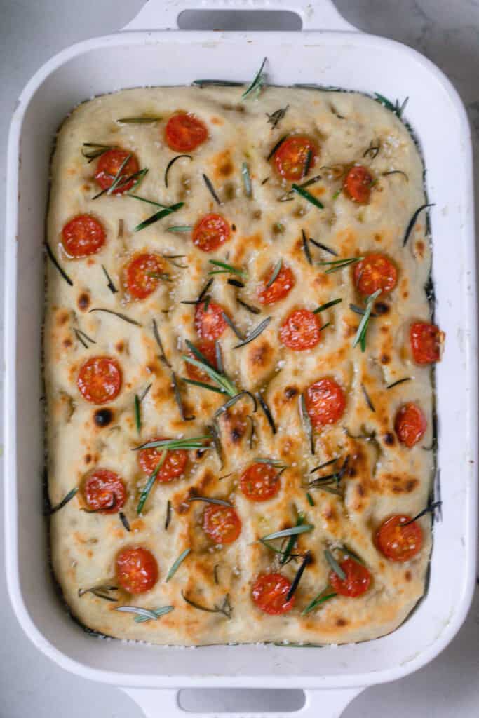 overhead photo of a white baking dish with a freshly baked sourdough focaccia bread topped with fresh rosemary and sliced tomatoes