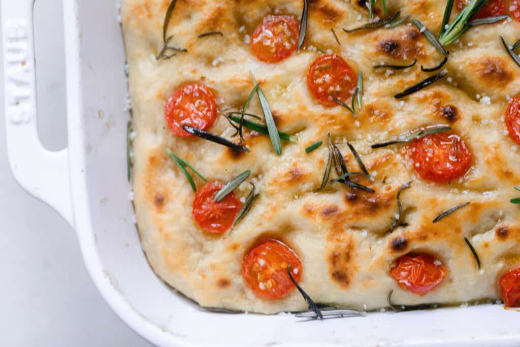 overhead photo of sourdough focaccia topped with fresh rosemary and slice tomatoes in a white baking dish