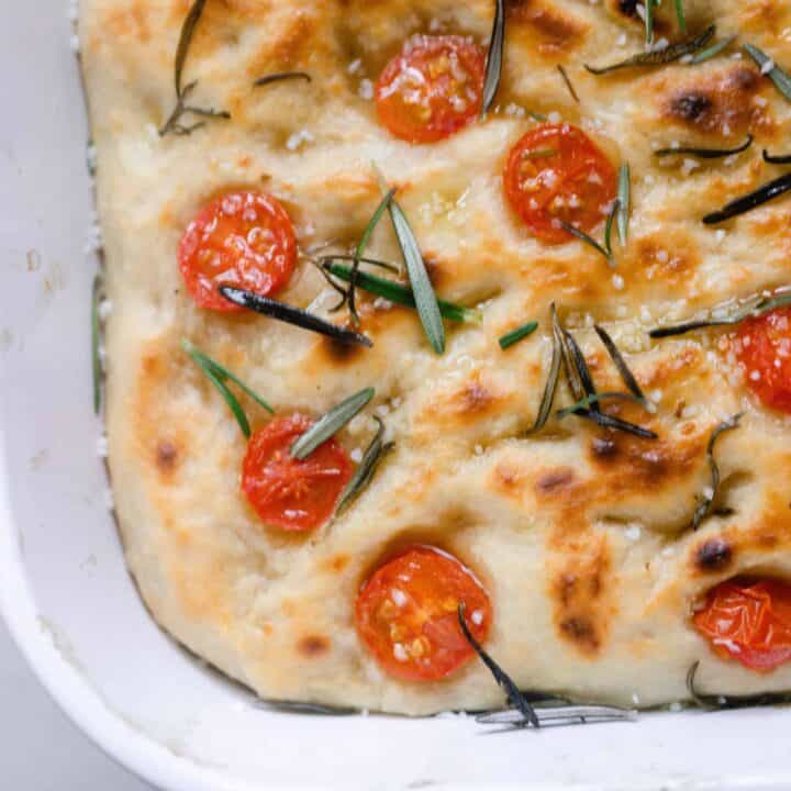overhead photo of sourdough focaccia topped with fresh rosemary and slice tomatoes in a white baking dish
