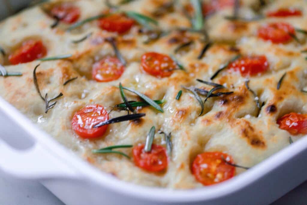 side view of sourdough focaccia in a white baking dish topped with tomatoes and rosemary