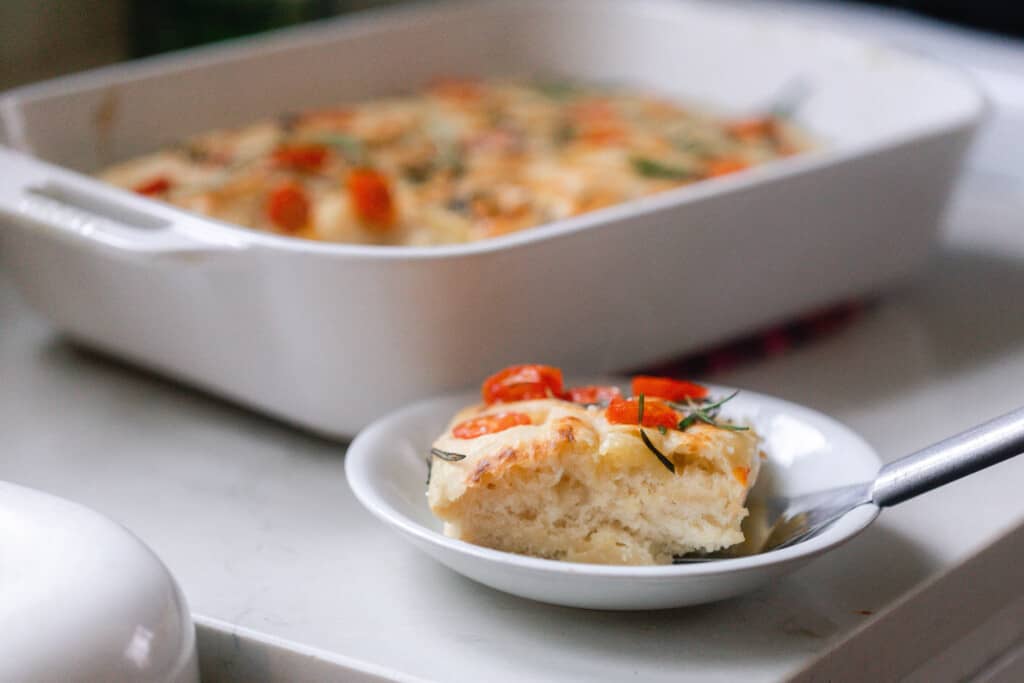 slice of sourdough focaccia bread on a white plate with a spatula. A White baking dish with more focaccia is behind the plate