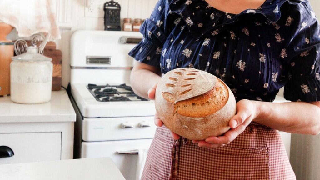 a lady wearing a blue shirt and red apron holding a loaf of bread in a white kitchen