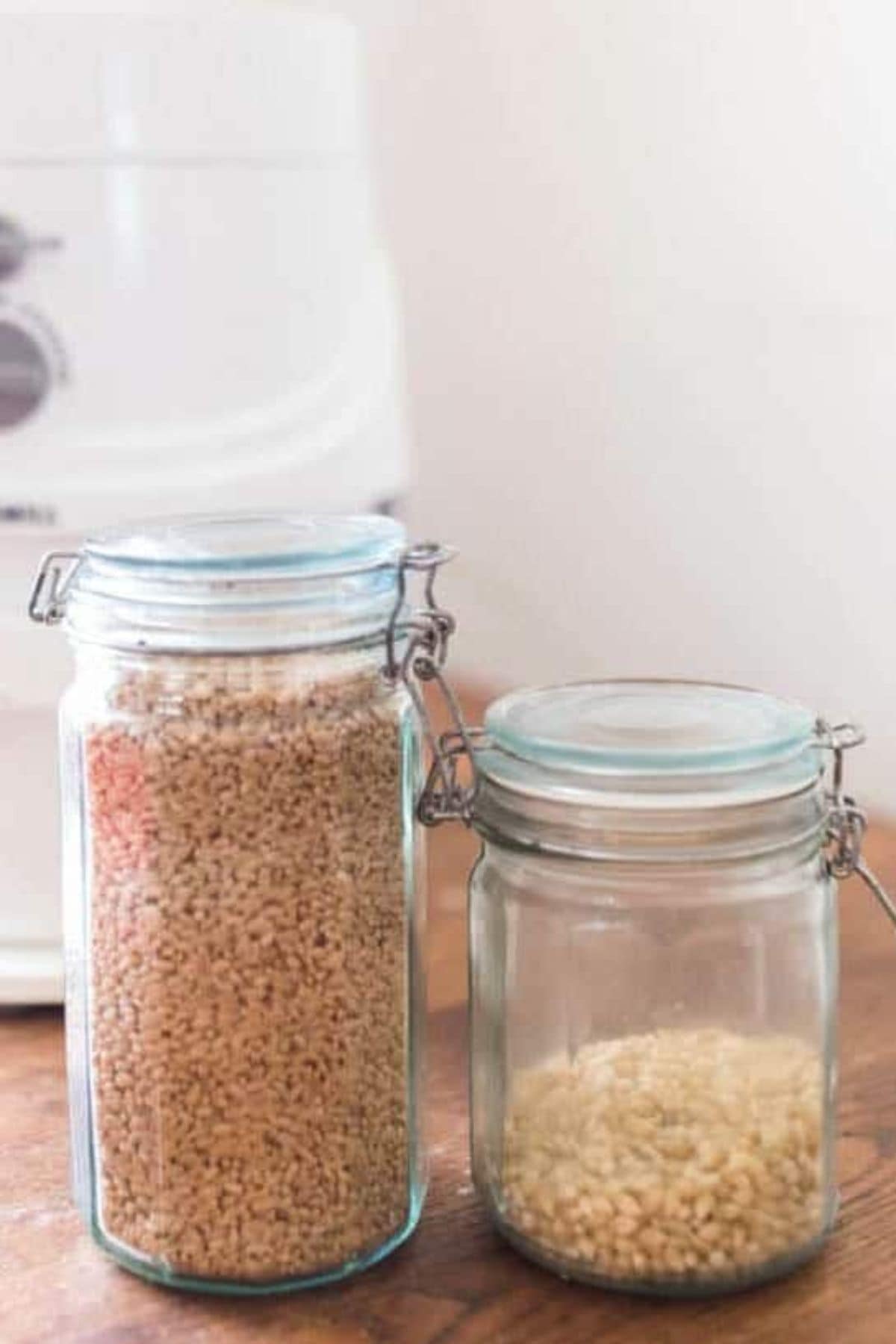 A large jar with wheat berries next to a small jar of popcorn kernels on a wooden counter. 