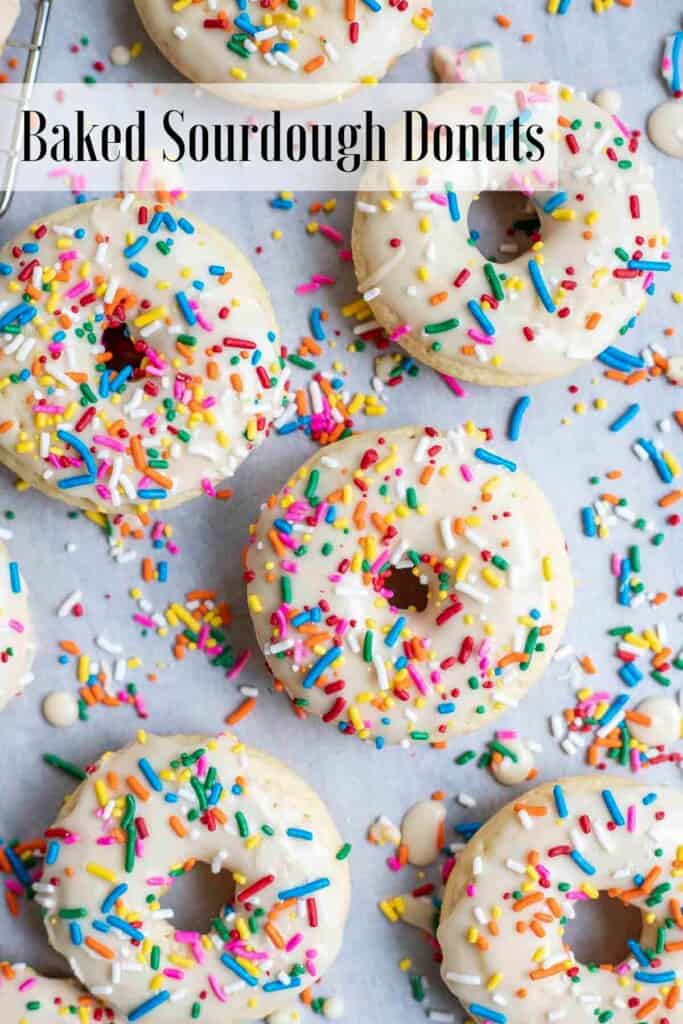 overhead photo of 6 baked sourdough donuts with vanilla frosting and sprinkles on parchment paper covered in sprinkles