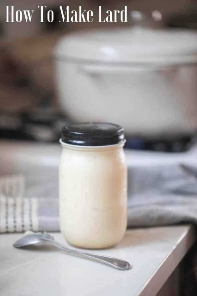 homemade lard in a glass mason jar with a black lid on a white countertop with a spoon in from of the jar. A white dutch oven is in the background