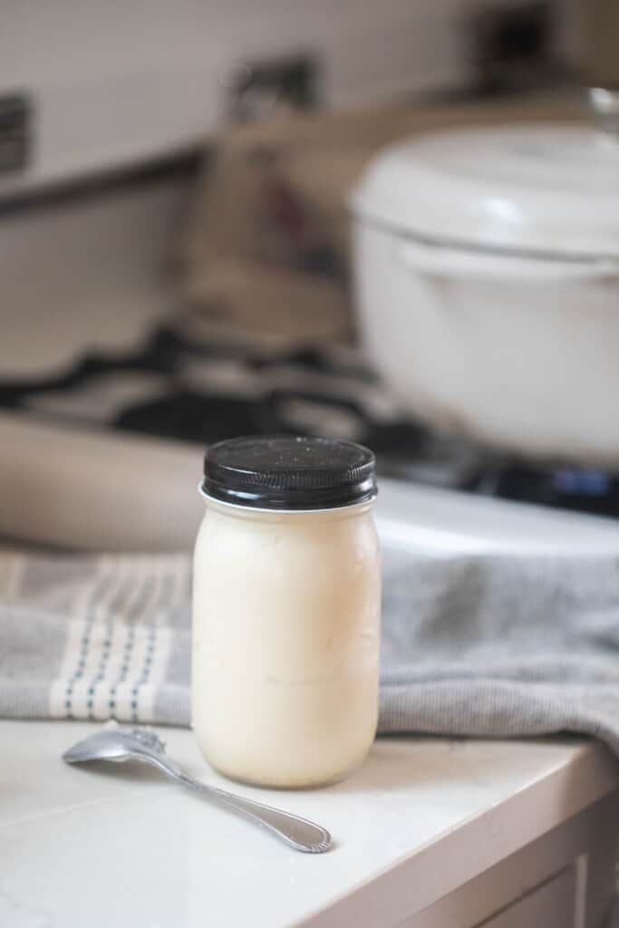 mason jar full of lard with a black lid on a white countertop. A silver spoon sits in front of the jar and a gray and white towel sit behind it.