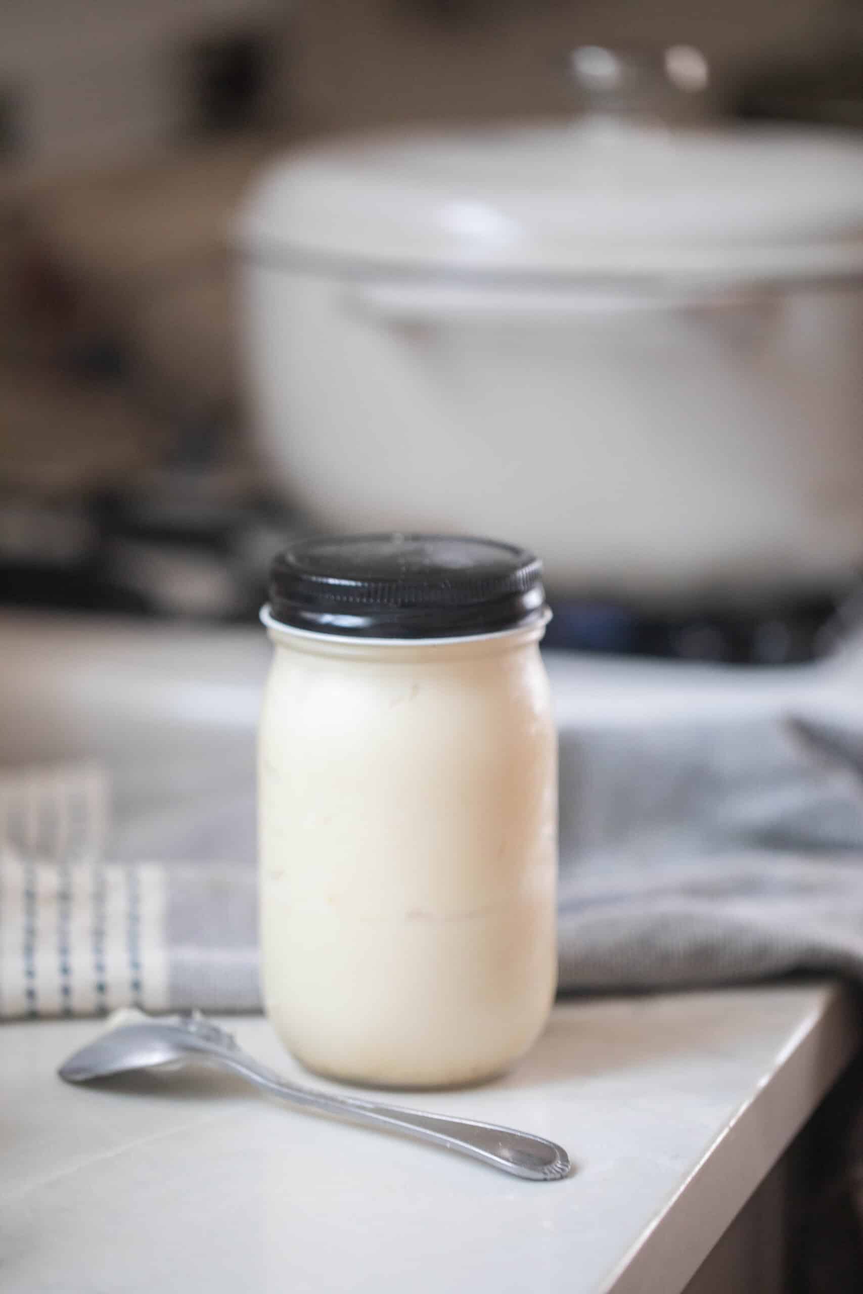 homemade lard in a glass mason jar with a black lid on a white countertop with a spoon in from of the jar. A white dutch oven is in the background