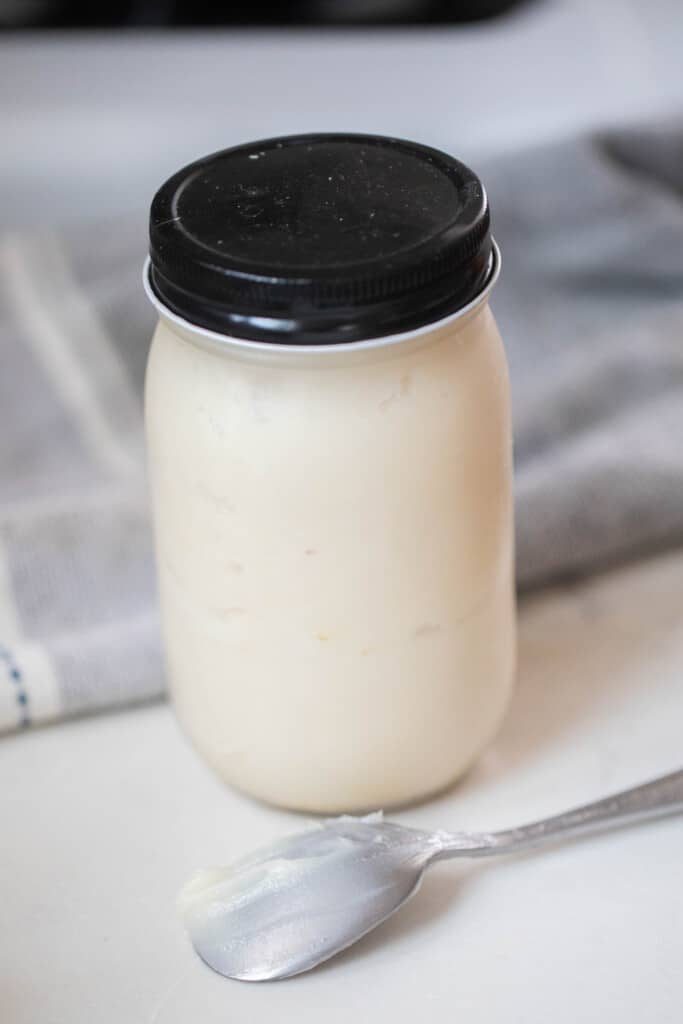 lard in a glass mason jar with a black lid on a white countertop with a metal spoon in the front and a blue and gray towel in the background