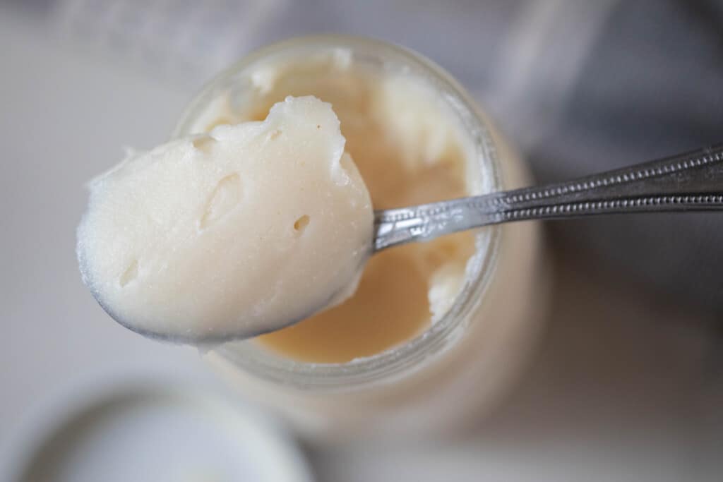 overhead photo of a spoonful of lard resting on the rim of a jar of lard. The jar is placed on a white countertop next to a towel