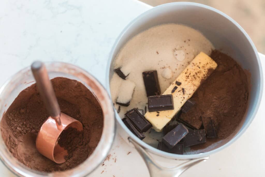 a pot with coco, butter, chocolate, and sugar next to a bowl of cocoa powder