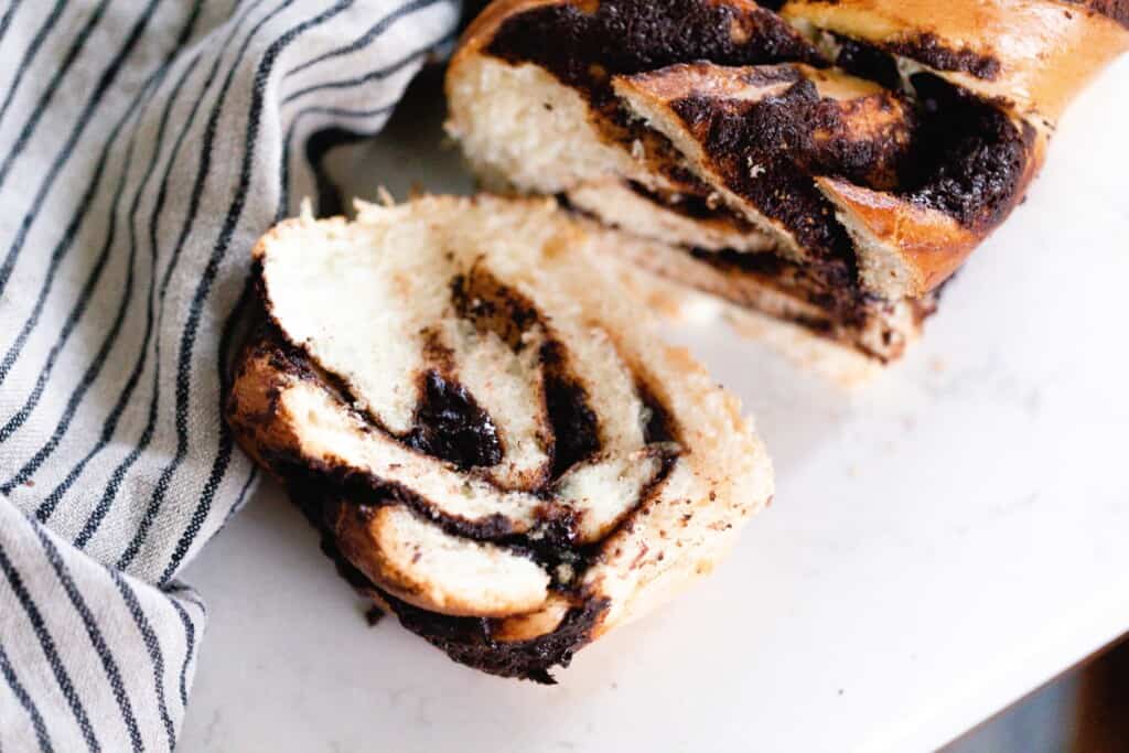 slice of sourdough chocolate babka sliced off of the loaf of bread on a white countertop with a black and cream stripped towel to the left