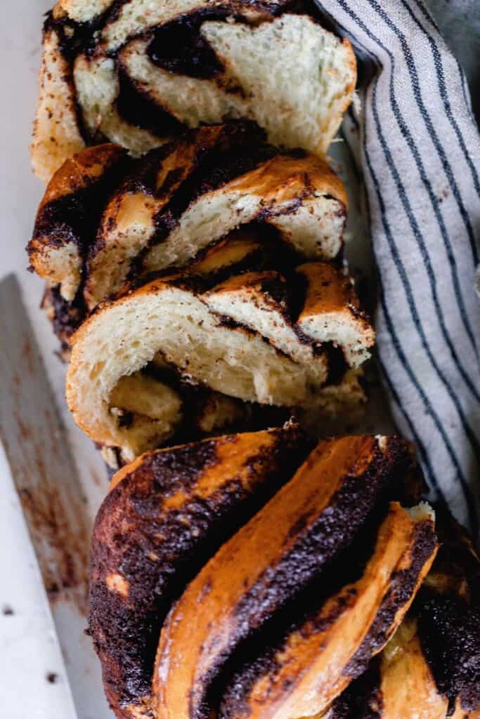 loaf of sourdough babka with swirls of chocolate filling  sliced on a white countertop with a black and cream stropped towel to the right.