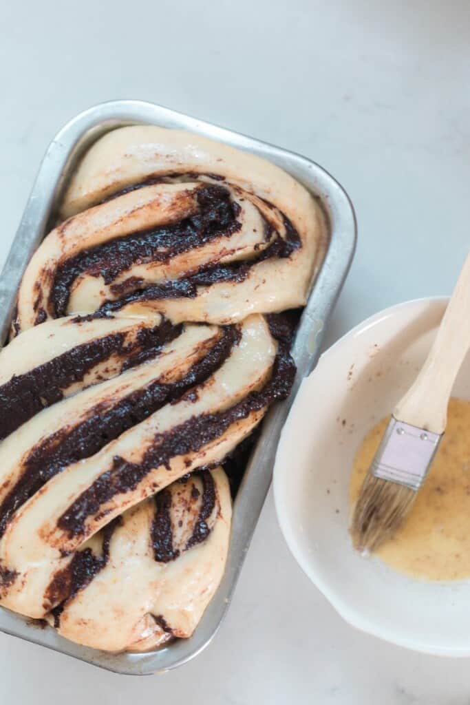 sourdough babka dough risen in a loaf pan and brushed with egg wash. A small bowl with an egg wash and pastry brush sit to the right of the dough