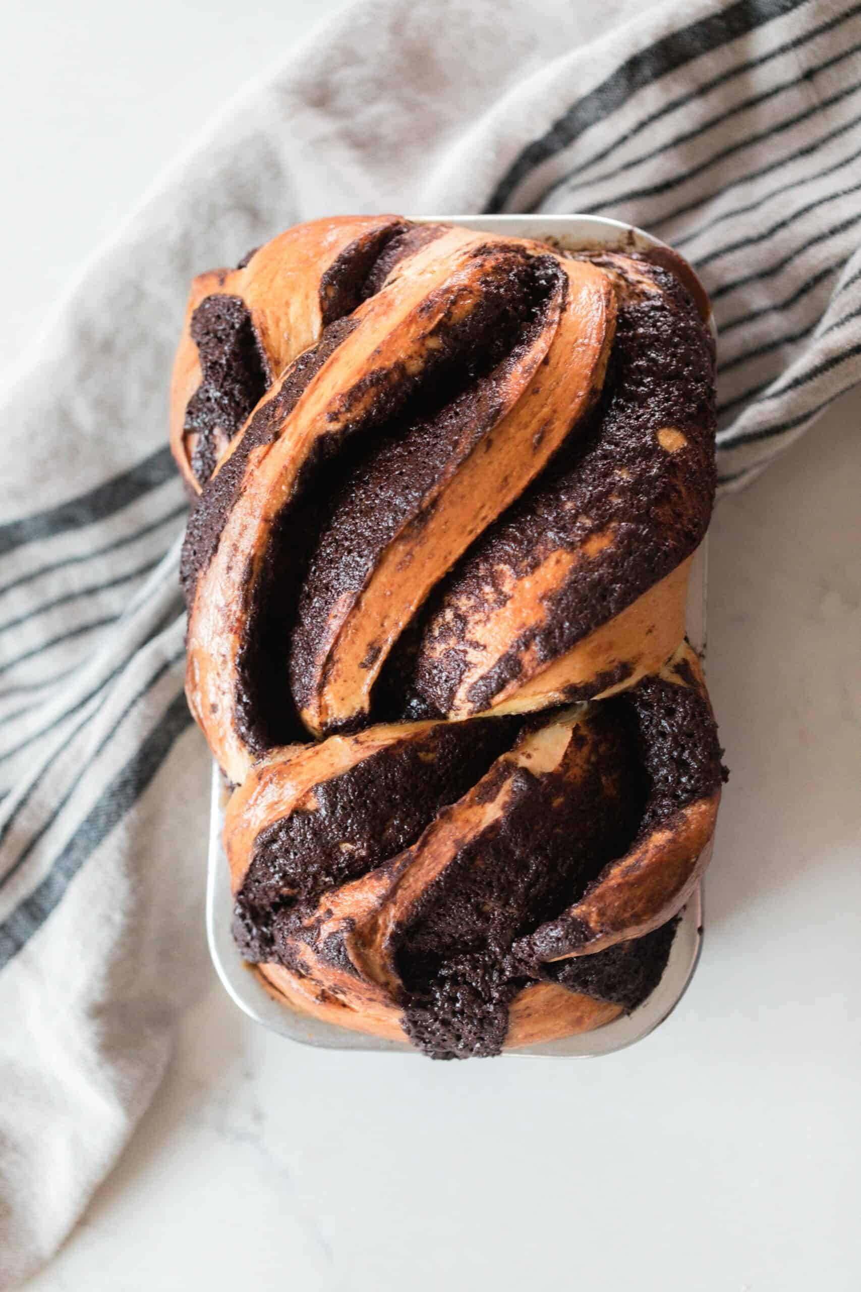 overhead photo of a loaf of sourdough chocolate babka on a white countertop and a black and cream colored stripped towel