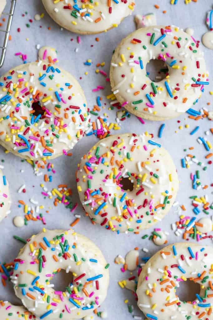 overhead photo of 6 baked sourdough donuts with vanilla frosting and sprinkles on parchment paper covered in sprinkles