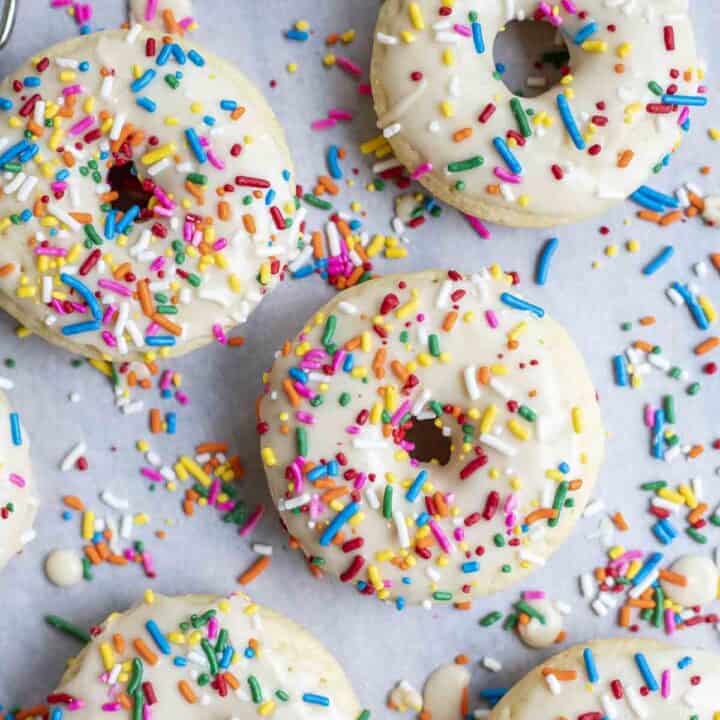 overhead photo of 6 baked sourdough donuts with vanilla frosting and sprinkles on parchment paper covered in sprinkles