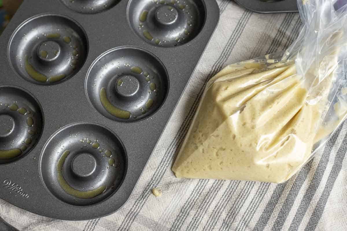A greased donut pan on a stripped towel with a plastic bag with donut filling to the right.