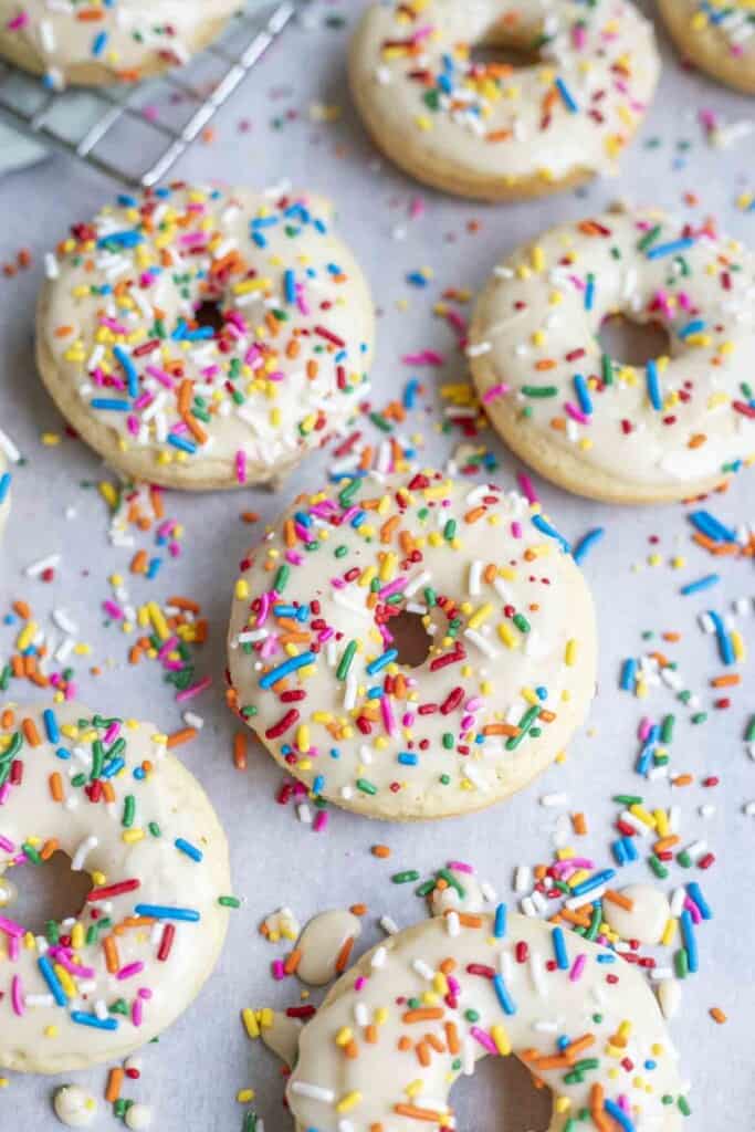 six baked sourdough donuts topped with vanilla glaze with sprinkles on parchment paper with more sprinkles surrounding the donuts