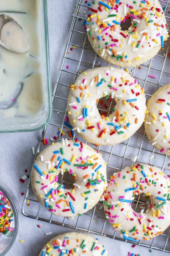 five sourdough baked donuts topped with vanilla frosting and sprinkles on a wire rack with a glass dish of more frosting to the left