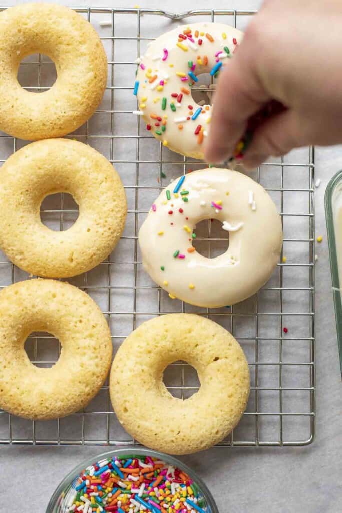 hand sprinkling sprinkles over a iced donut on a wire rack surrounded by more donuts