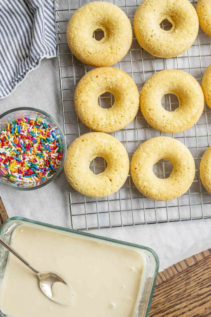 sourdough baked donuts fresh from the oven on a wire rack over parchment paper. A small jar to the left contains rainbow sprinkles and a rectangle dish of icing is in front.