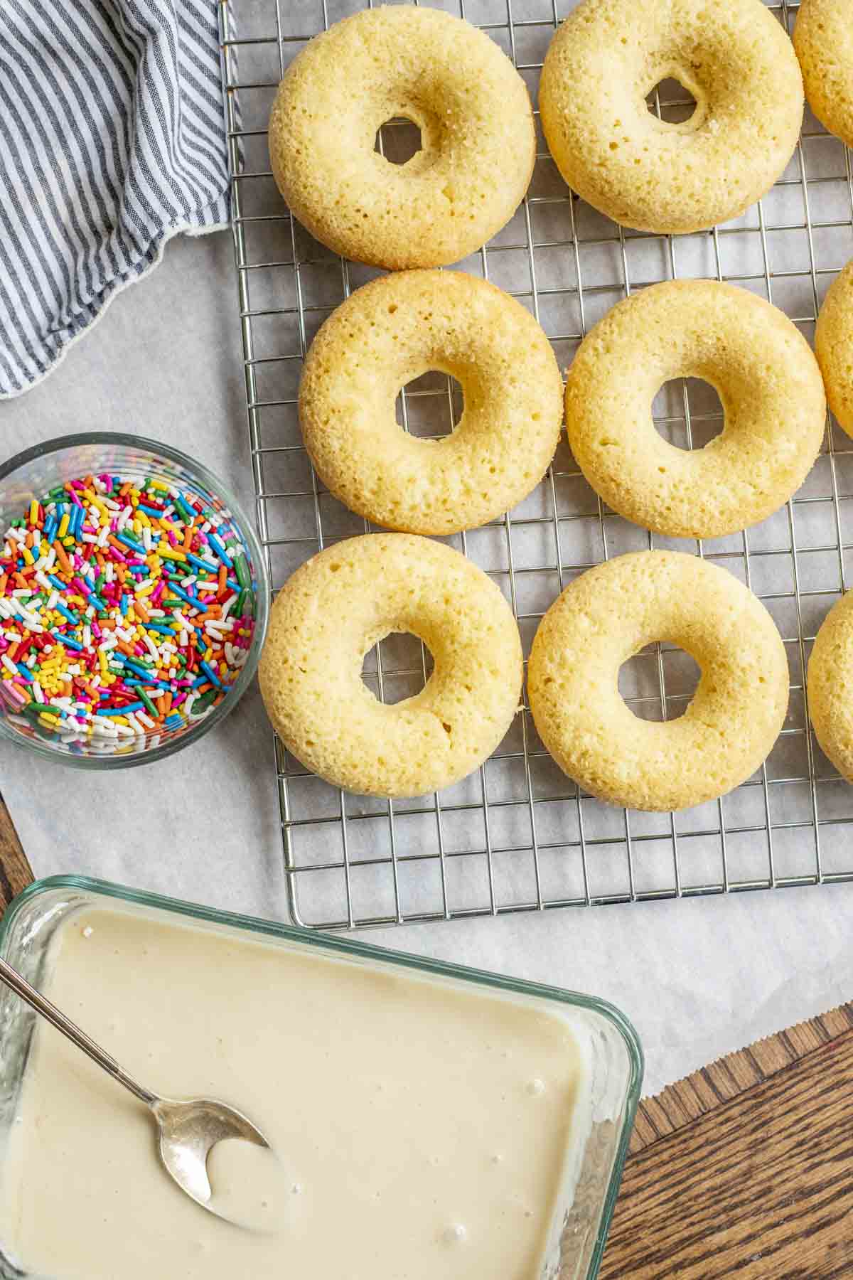 Freshly baked donuts on a wire rack with a bowl of glaze and a bowl of sprinkles next to it. 