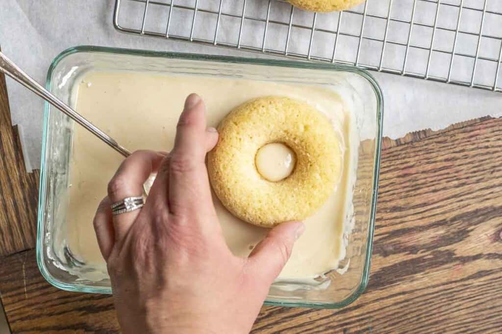 hand dunking a sourdough baked donut into a rectangular glass dish on a wood table next to a wire rack over parchment paper