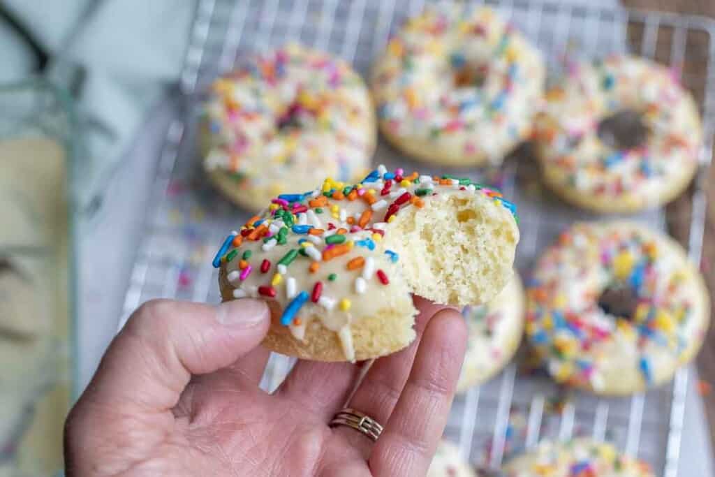 hand holding. a sourdough baked donut topped with vanilla frosting and sprinkles with a bite taken out. More donuts are in the background