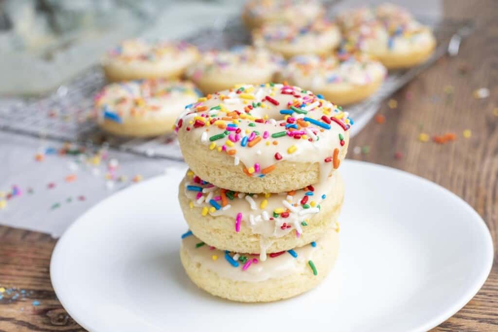 three baked sourdough donuts with vanilla frosting and sprinkles stacked up on a white plate on a wood table