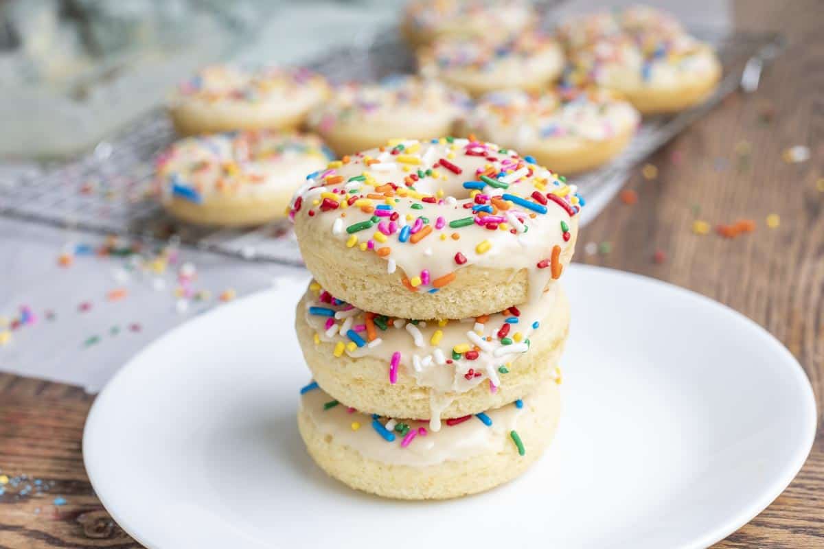 Three baked sourdough donuts with vanilla frosting and sprinkles stacked up on a white plate on a wood table.