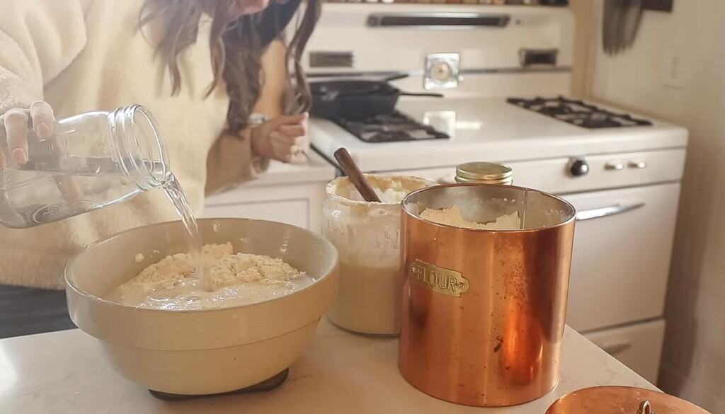 woman adding water to a bowl of flour on a kitchen scale. The bowl sits on a white countertop with a copped canister to the right
